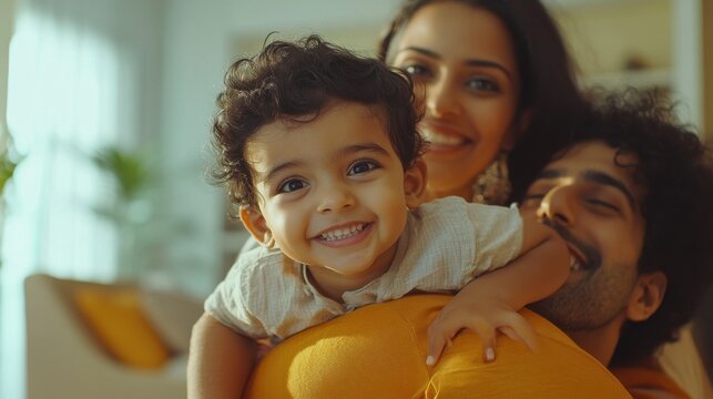 Cheerful african mother and indian father playing with son at home. Cute boy enjoying sitting on father shoulder while looking at camera. Middle eastern family having fun together on the sofa at home