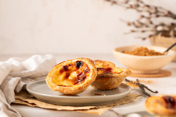 Two Portuguese custard tarts, pastel de nata, with caramelized tops on a gray plate, placed on a cork mat on a light surface, accompanied by a small branch with buds