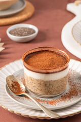 Tiramisu overnight oats and chia pudding topped with creamy yogurt and cocoa powder, served in a glass jar on a white plate with a spoon sprinkled with cocoa powder