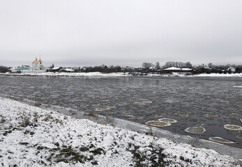 View of a freezing river. Round ice floes are forming in the water. Ice disks are smoothly rotating on the water surface. The grass is covered with fresh white snow. A cloudy winter day. 