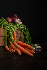 View of a bunch of carrots with green leaves, onions and garlic on a wooden box, black background, vertical with copy space