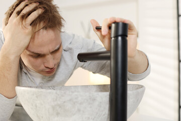 Unhappy young man suffering from hangover near sink in bathroom
