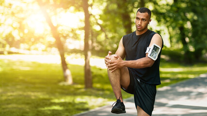 Handsome black athlete stretching his leg after jogging at summer park, panorama