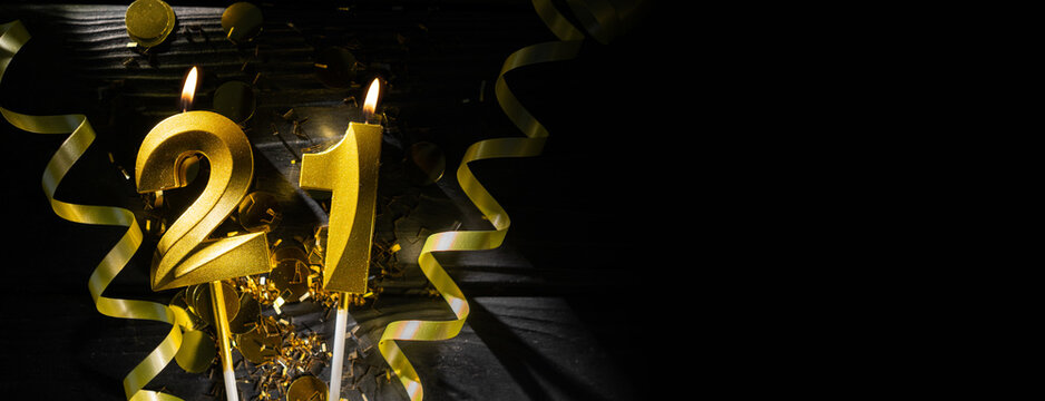 Golden candle shaped as number 21 burns on dark table, surrounded by shimmering confetti and curling ribbons, creating festive atmosphere. Concept of celebrating milestone birthday