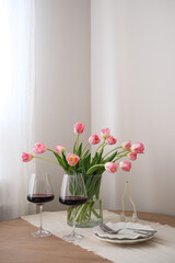 A beautiful bouquet of pink tulips beside a glasses of rich red wine, candles, plates enhance home decor and ambiance. Table serving.