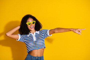 Young cheerful woman with curly hair pointing and smiling, wearing striped shirt and yellow...