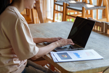 Close-up of Asian woman working on laptop with business reports on wooden table. Hands typing on...