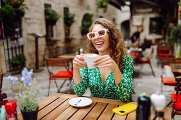 Cheerful woman in stylish green dress enjoying morning coffee in cozy cafe. Young tourist drinking hot drink at wooden table outdoors. Drink, tourist and fun concept.