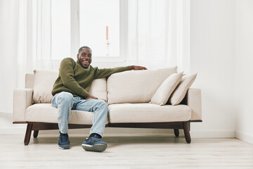 Obraz premium Happy Black man in cozy sweater on beige couch, showcasing comfort and relaxation in a bright, modern living room environment