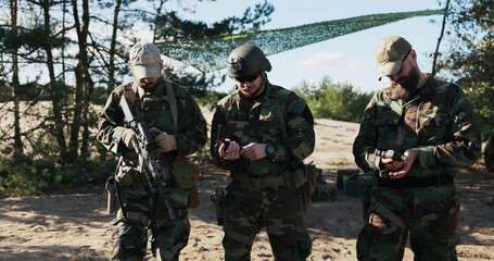 A team of three soldiers in camouflage stands in readiness, inspecting weapons, unlocking pistols, adjusting rifles, and ensuring all gear is secured in holsters and belts.