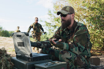 Fototapeta premium Soldiers in full combat gear wait in the field for orders while their commander, sitting at a computer outside, engages in an online video call with the boss.