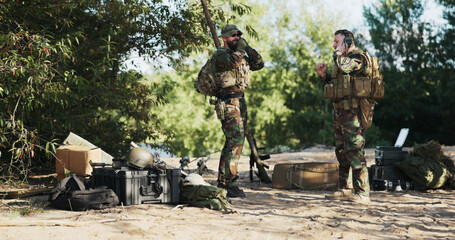 Fototapeta premium Two soldiers in camouflage uniforms prepare for field exercises, securing heavy backpacks, ammo, and weapon belts, ready for a territorial defense mission near the base in the bushes.