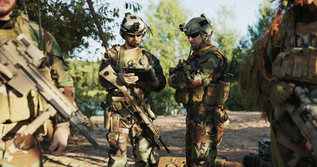 Fototapeta premium Soldiers in camouflage uniforms with rifles over their shoulders and helmets on their heads check mission details on a tablet while contacting the army base before field exercises.