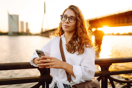 Cute woman walks along the embankment at sunset with a phone in her hands. A beautiful woman with a phone chats outdoors. Blogging, leisure concept.