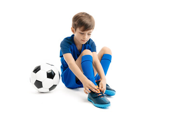 Boy soccer player in blue sport outfit posing sitting on the floor on white background. Boy is tying his shoelaces