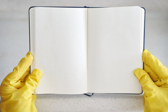 Open blank dot grid notebook held by person wearing yellow cleaning gloves on light surface. - Powered by Adobe