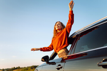 Young woman feels freedom leaning out of car window. Beautiful female traveler enjoying sunset from car. Car travel, leisure concept. Active lifestyle. © maxbelchenko