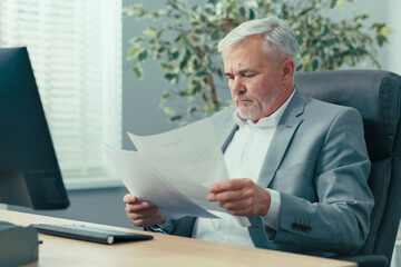 An experienced businessman, grey-haired man head of company is sitting at desk in a comfortable...