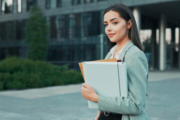 Fototapeta premium A smiling, pretty, successful girl running her own company manager boss is standing next to glass modern building going to meeting conference to work holding files with documents in her hand