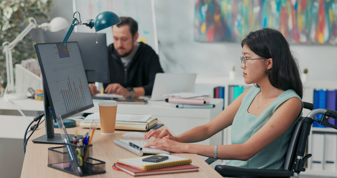 Workday in office, corporation, company, co-workers are working on computers, drafting documents, woman in glasses with Asian beauty is sitting at desk in wheelchair, she is disabled, after accident