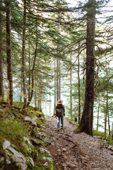 Obraz premium A young tourist with a hiking backpack walks along a hiking path in a mountain forest. Happy woman enjoys the weather outdoors. Hiking, travel concept. Active lifestyle.