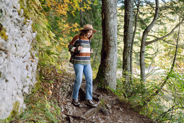Naklejka premium A young tourist with a hiking backpack walks along a hiking path in a mountain forest. Happy woman enjoys the weather outdoors. Hiking, travel concept. Active lifestyle.