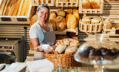 Smiling female baker holding a wicker basket brimming with freshly baked bread stands in front of shelves displaying a variety of artisan loaves in a charming bakery shop