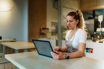 Female baker wearing apron using laptop, managing online orders and inventory in modern bakery shop, ensuring efficient service and customer satisfaction