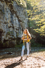 Naklejka premium Young woman with a hiking yellow backpack hiking in the mountains near a mountain river. Happy female traveler enjoying sunny weather outdoors. Travel concept, hiking. Active lifestyle.