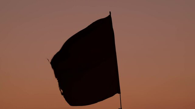 Waving red flag against a stunning sunset backdrop. Red flag waves in the wind during sunset. The sun sets behind the flag, casting a warm, pink glow across the sky. Red and yellow flag on a beach