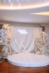 Elegant wedding stage with white drapery, floral arrangements, and neon sign. Luxurious reception decor with a round dance floor and crystal chandeliers