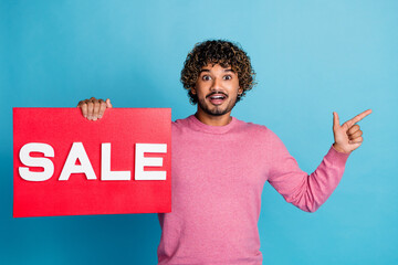 Excited young man in pink sweater holds red sale sign and points enthusiastically against blue...