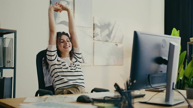 Beautiful Caucasian woman stretching arms above head while sitting in office chair. Enjoying break after long work session. Finishing important assignment. Feeling relaxed and reenergized.