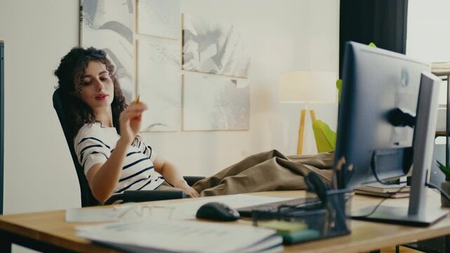 Beautiful Caucasian woman leaning back in office chair while holding pencil and smiling. Resting feet on wooden desk and looking at computer screen. Thinking about work or daydreaming.