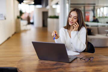Cheerful young beautiful woman talking on mobile phone and using laptop with smile while sitting at her working place