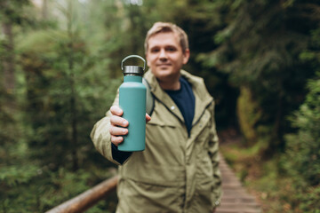 Happy blonde man in sport clothes having a halt after hiking. 30s Hiker drinking water from water bottle or hot drink from blue thermos. Travel, adventure, active lifestyle. High quality photo.