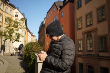 Side view of young adult bearded man wearing sunglasses, hat and black puffer jacket using using...