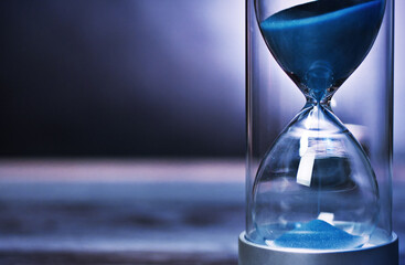 Blue sand flowing through hourglass measuring time passing by. A dark background with copy space