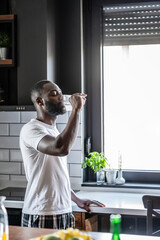 An African American man in a white t-shirt and plaid pajama pants drinks a glass of water in a...