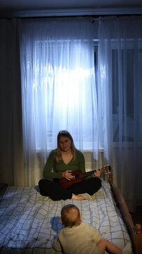 A young woman strums a ukulele while sitting cross-legged on a bed, softly illuminated by natural light. A child looks on with curiosity, capturing a peaceful domestic scene.