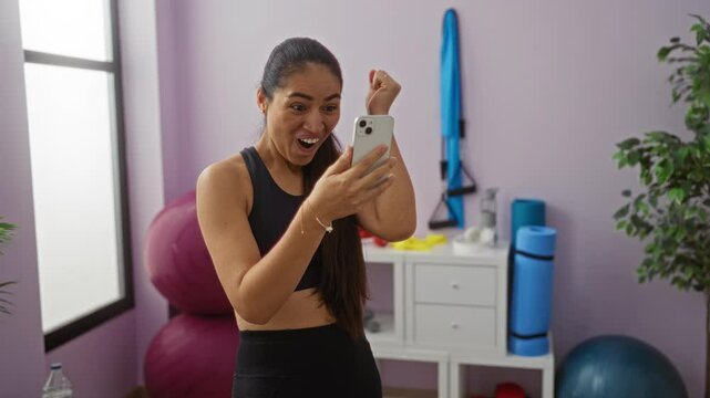 Smiling woman in gym holding phone looking excited in sports environment with exercise equipment visible in background.