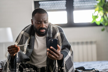 Sick man with a plaid blanket, wearing pajamas, sitting on a bed, checking his phone while holding...
