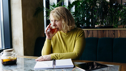 A senior businesswoman enjoys a beverage while using a tablet, embracing the freelance remote work concept. Her relaxed demeanor in a modern workspace reflects the flexibility and freedom of digital e