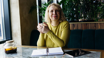 A senior businesswoman enjoys a beverage while using a tablet, embracing the freelance remote work concept. Her relaxed demeanor in a modern workspace