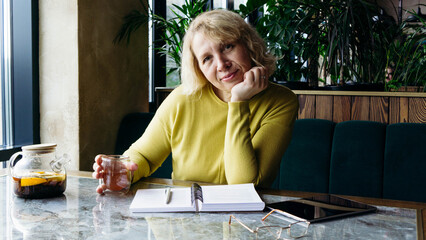 A senior businesswoman enjoys a beverage while using a tablet, embracing the freelance remote work concept. Her relaxed demeanor in a modern workspace