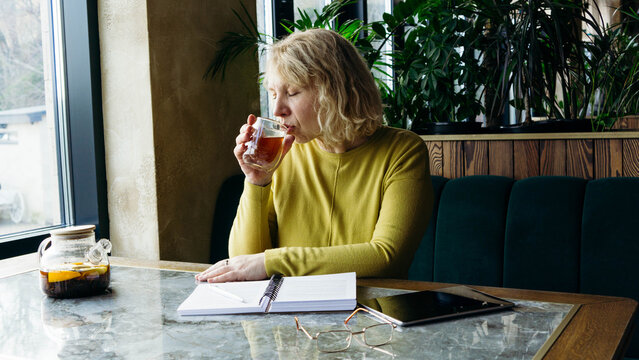 A senior businesswoman enjoys a beverage while using a tablet, embracing the freelance remote work concept. Her relaxed demeanor in a modern workspace - Powered by Adobe