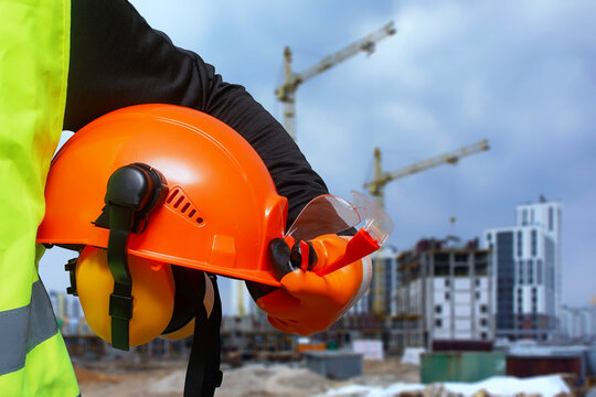 Construction worker in a high-visibility vest holding an orange safety helmet and protective gear at a building site.
