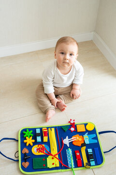 Baby girl plays on the floor with a developing bright handmade toy for fine motor skills made of felt. Early childhood development, soft busy book with with laces, zippers and Velcro