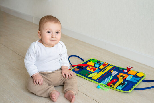 Cute baby girl smile on the floor. On the background developing bright handmade toy for fine motor skills made of felt. Early childhood development, soft busy book with with laces, zippers and Velcro.