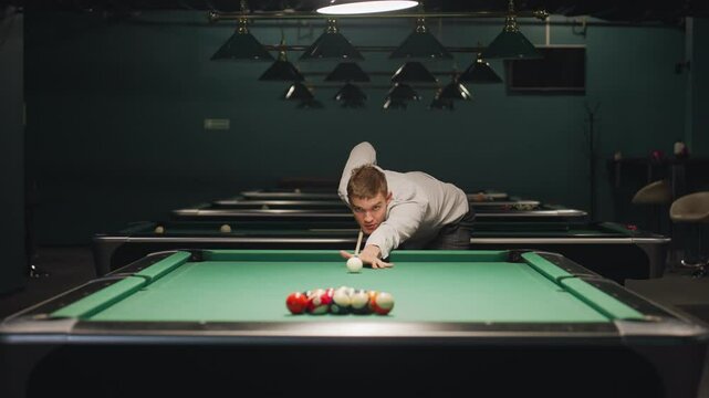 Billiard player in white shirt and grey trousers leans over green pool table, gripping cue stick with intense focus. Dark billiard room enhances concentration. Overhead lights